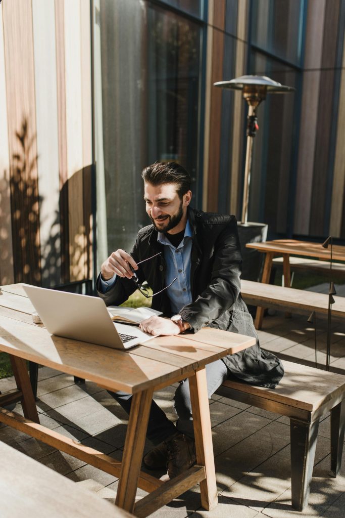 Man sitting at a partly shaded picnic table smiling and working on this computer.